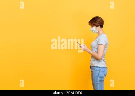 Jeune femme asiatique portant un masque médical debout et utilisant téléphone portable isolé sur fond jaune studio avec espace de copie Banque D'Images