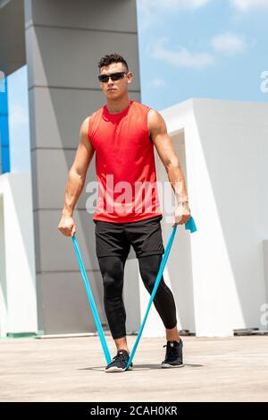 Beau sportif faisant l'exercice d'élévation latérale de l'épaule avec la bande de résistance à l'extérieur, sur le toit, au soleil Banque D'Images