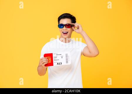 Portrait à la taille du jeune homme asiatique heureux portant la 3D lunettes tenant un billet de cinéma isolé sur fond jaune de studio Banque D'Images