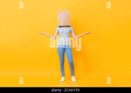 Anonyme femme en tenue décontractée tête de couverture avec sac en papier faire un geste de main ouverte sur fond jaune de studio Banque D'Images