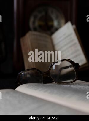 Vieilles lunettes usées reposant sur un livre ouvert avec des livres et une horloge en arrière-plan. Notion : temps de lecture Banque D'Images