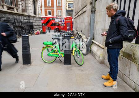 LONDRES - 04 FÉVRIER 2020 : un homme qui exploite un smartphone et regarde un vélo de location d'assistance électrique Lime. Bus Red London Double Decker à l'entrée Banque D'Images