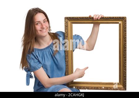 Une jeune fille avec de longs cheveux dans une robe bleue. Isolé sur un fond blanc, tenant un cadre d'image dans ses mains, pointant vers lui avec son doigt. copyspace. Avec une expression heureuse et souriante sur son visage Banque D'Images