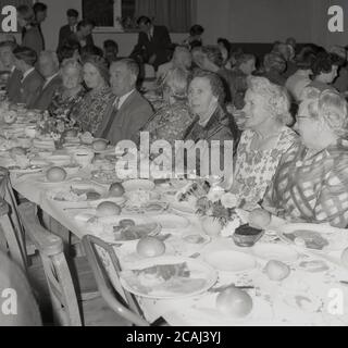 1964, historique, un groupe de retraités et de personnes locales assis à un souper de récolte dans la salle du village à Walton, Aylesbury, Buckinghamshire, Angleterre, Royaume-Uni. À cette époque, les grands repas pour célébrer la récolte étaient communs dans les zones rurales de l'Angleterre, auxquelles tous les participants à la récolte ont été invités. L'idée d'un repas partagé par toute la communauté pour vous remercier pour une récolte réussie est une tradition très ancienne et profondément assise. Le repas a normalement eu lieu le jour de Michaelmas. Banque D'Images