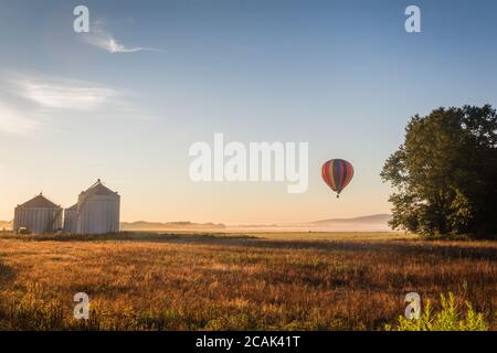 Le ballon d'air chaud flotte sur le champ de ferme rempli de brouillard et collines ondulantes au lever du soleil Banque D'Images