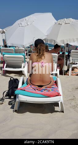 La jeune femme du bikini est assise sur une chaise longue et des bains de soleil sur une plage de luxe avec des parasols blancs sur fond bleu ciel. Vacances d'été en mer Banque D'Images