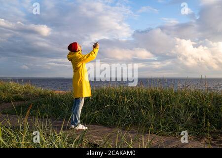 Un homme en imperméable jaune porte un chapeau rouge sur la plage, sur un chemin en bois, regarde un ciel nuageux et prend des photos sur un smartphone. Saison d'automne. Banque D'Images