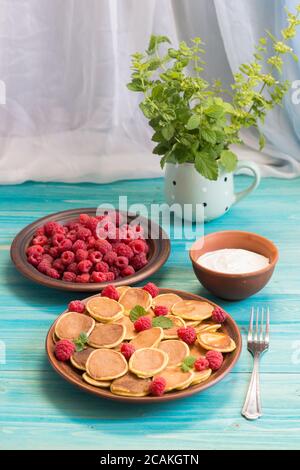 Céréales pancakes et framboises fraîches mûres rouges dans une assiette brune. Un bouquet de Melissa dans une tasse sur une table en bois bleu près de la fenêtre. Un été délicieux Banque D'Images