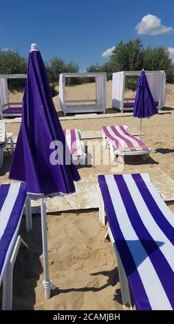 Parasol fermé près d'une chaise longue blanche à rayures bleues et de parasols carrés blancs sur du sable de plage propre avec des nuages blancs dans le ciel bleu. Jour ensoleillé a Banque D'Images