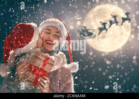 Joyeux Noël et joyeuses fêtes ! Maman et fille échangent des cadeaux à l'extérieur. Nuit de Noël. Le Père Noël vole dans son traîneau contre le ciel lunaire. Por Banque D'Images