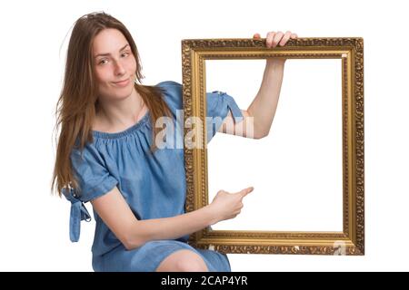 Une jeune fille avec de longs cheveux dans une robe bleue. Isolé sur un fond blanc, tenant un cadre d'image dans ses mains, pointant vers lui avec son doigt. copyspace. Avec un regard malicieux Banque D'Images