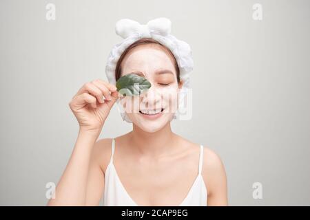 Femme asiatique dans une serviette appliquant un masque blanc d'argile faciale et tenant une feuille. Banque D'Images