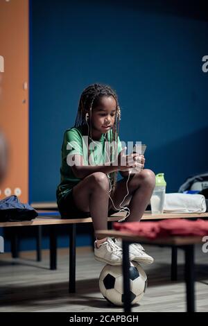 Un petit joueur de football apprécie le contenu d'un téléphone cellulaire dans un vestiaire Banque D'Images