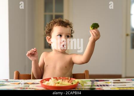 Une adorable petite fille assise à la table aime manger des légumes directement à la main. Banque D'Images