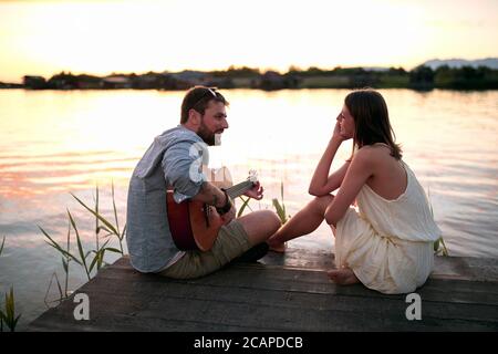 magnifique femme caucasienne appréciant en compagnie d'un homme beardy jouant de la guitare au bord du lac au coucher du soleil Banque D'Images