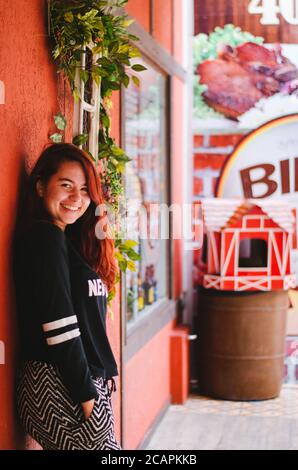 Un jeune adulte redhead brésilien souriant à l'appareil-photo pendant poser à côté d'un magasin exposé avec des murs rouges et une poubelle en forme de joli colonel rouge Banque D'Images