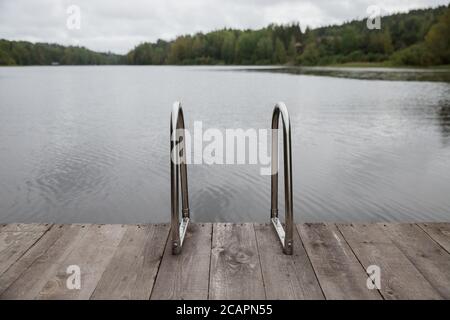 Main courante en métal pour la descente dans le lac, échelle en fer pour les nageurs. Jetée en bois sur le lac, ciel nuageux et eau calme du lac en arrière-plan. Banque D'Images