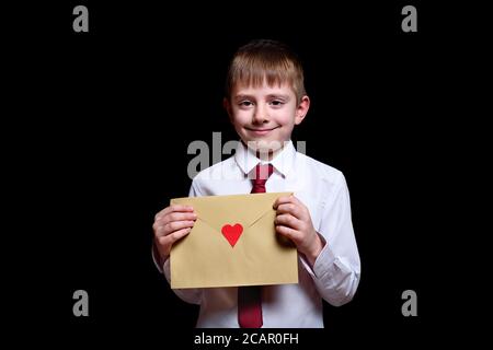 Un garçon aux cheveux justes dans une chemise et une cravate tient une enveloppe avec un coeur. Isoler sur fond noir Banque D'Images