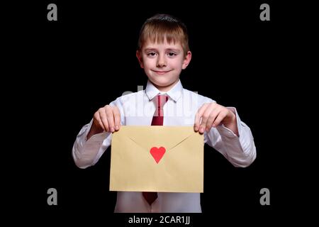 Un garçon aux cheveux justes dans une chemise et une cravate tient une enveloppe avec un coeur. Isoler sur fond noir Banque D'Images