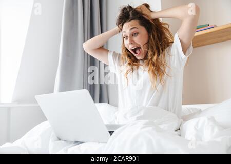 Portrait d'une femme excitée au gingembre exprimant sa surprise et utilisant un ordinateur portable assis dans le lit après le sommeil Banque D'Images