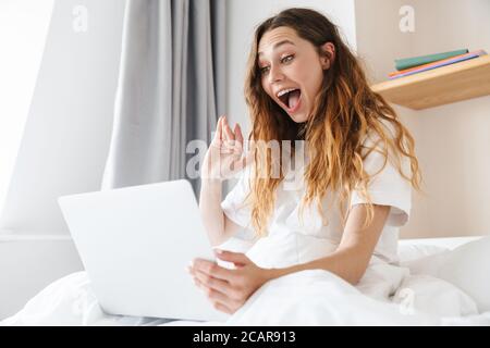 Portrait d'une femme excitée au gingembre en agitant la main et en utilisant un ordinateur portable assis dans le lit après le sommeil Banque D'Images