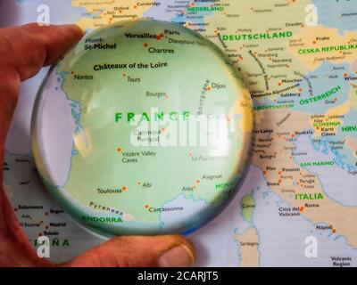 Gros plan POV vue aérienne d’un doigt et d’un pouce tenant une loupe à dôme sur une carte de l’Europe, avec la France agrandie pour une vue plus rapprochée. Banque D'Images