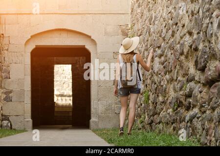 Fille dans un chapeau et avec un sac à dos marchant le long de la forteresse, la lumière du soleil. Vue arrière Banque D'Images