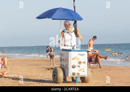 Punta Umbria, Huelva, Espagne - 7 août 2020: Une vendeuse de rue féminine vendant de la glace sur la plage. Le vendeur porte un masque de protection ou un masque médical Banque D'Images