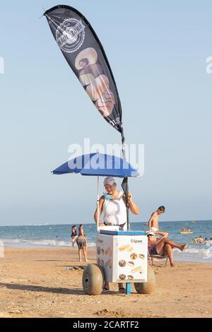 Punta Umbria, Huelva, Espagne - 7 août 2020: Une vendeuse de rue féminine vendant de la glace sur la plage. Le vendeur porte un masque de protection ou un masque médical Banque D'Images