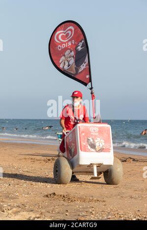 Punta Umbria, Huelva, Espagne - 7 août 2020: Une vendeuse de rue féminine vendant de la glace sur la plage. Le vendeur porte un masque de protection ou un masque médical Banque D'Images