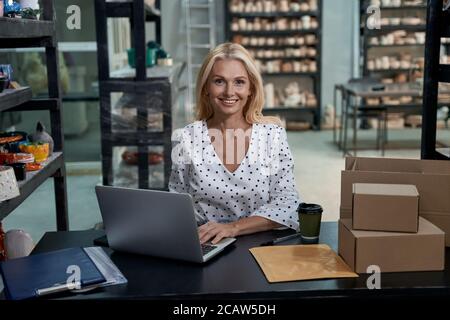 Vente en ligne. Une femme heureuse propriétaire d'affaires ou une femme d'affaires assise dans son studio d'art ou dans une boutique de poterie artisanale avec des produits faits à la main en céramique, travaillant sur un ordinateur portable et regardant l'appareil photo. Petite entreprise Banque D'Images
