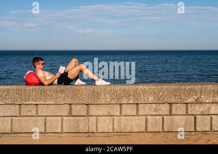 Un jeune homme se pose sur un mur inférieur pendant la lecture un livre avec la mer bleue en arrière-plan dans Scarborough Banque D'Images