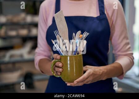 Création du processus. Femme d'artisanat travaillant dans son atelier. Gros plan de mains de femmes tenant un bol en céramique rempli de brosses et d'autres outils. Boutique de poterie artisanale, petite entreprise Banque D'Images