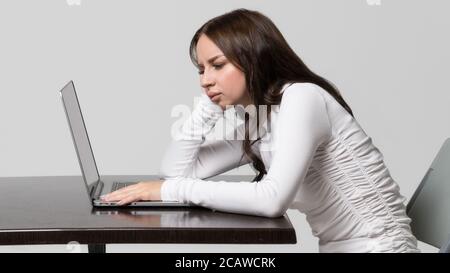 Mauvaise posture, position de posture altérée, défaut scoliose, concept orthopédique. Jeune femme assise à la table, regardant et travaillant à l'ordinateur portable, isolée. RA Banque D'Images