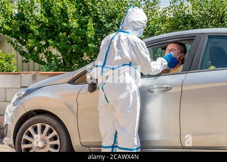 Un professionnel de la santé entièrement couvert d'un costume de protection blanc et de lunettes fait fonctionner un écouvillon pour Covid-19 à un homme assis à l'intérieur de sa voiture Banque D'Images