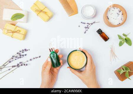 Les femmes tiennent les produits cosmétiques biologiques près du savon naturel, du sel de bain et des fleurs de lavande sur une table blanche, vue du dessus. Banque D'Images