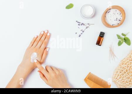 Vue de dessus des mains de femmes appliquant de la crème cosmétique à côté des produits de beauté naturels et des accessoires de bain sur la table blanche. Banque D'Images