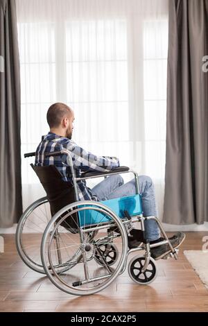 Inquiet jeune homme en fauteuil roulant vivant seul sérieux triste homme caucasien portant des vêtements décontractés et regarder la grande vue panoramique dans le salon moderne lumineux, l'hôpital ou la clinique. Banque D'Images