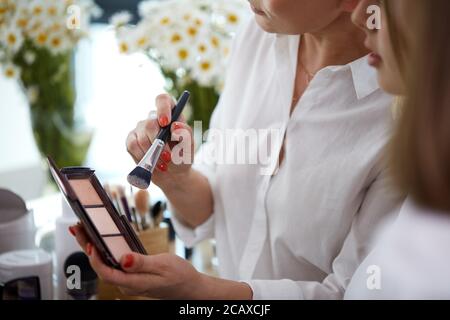 jeune artiste de maquillage caucasien qui va appliquer de la rougissement sur le visage du modèle, elle utilise des cosmétiques décoratifs professionnels de suring travail Banque D'Images