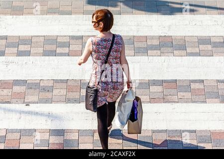 Une femme vêtue d'une robe d'été portant des sacs de shopping traversant la route sur une traversée de zèbre, Londres, Royaume-Uni Banque D'Images