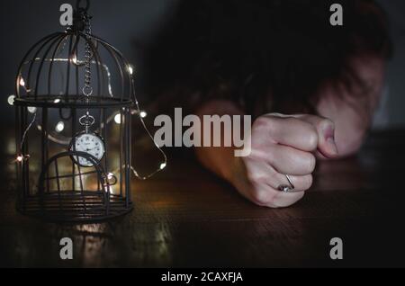 Photo conceptuelle de l'emprisonnement pendant le confinement du coronavirus. Une horloge emprisonnée dans une cage à oiseaux avec une femme blanche dépressive couché sur une table Banque D'Images