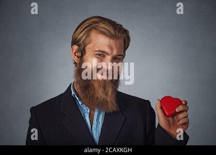 Les jeunes professionnels Caucasian businessman holding hipster barbu en forme de coeur rouge boîte-cadeau prêt pour la Saint-Valentin. Isolé sur mur gris gris studio Backg Banque D'Images