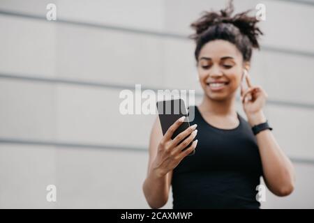 Musique et course à l'extérieur. Happy girl dans les vêtements de sport régulent les écouteurs sans fil et regarde le smartphone Banque D'Images