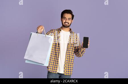 Achats en ligne. Homme indien tenant des sacs en papier et téléphone portable avec écran vide sur fond lilas, espace pour le design Banque D'Images