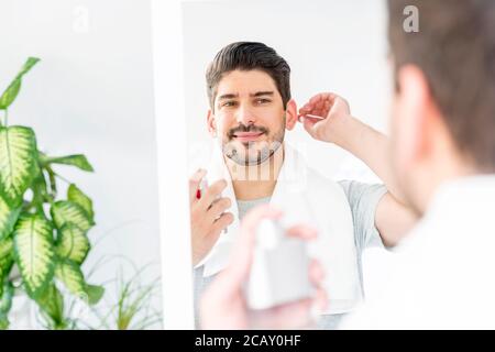 Beau homme appliquant de la lotion après-rasage sur le visage et souriant en se tenant au miroir de salle de bains. Banque D'Images