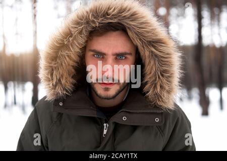 Beau homme sérieux et brutal barbu jeune caucasien debout à l'extérieur de la capuche en fourrure dans la forêt de saison d'hiver. Un sympathique gars européen qui marche en neige Banque D'Images