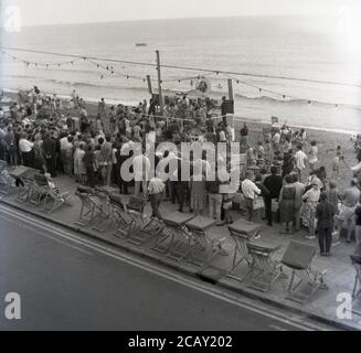 Années 1950, historiques, les gens en bord de mer qui regardent les vacanciers participer à un cycle à roulettes, sur une plate-forme spécialement construite sur une plage, Angleterre, Royaume-Uni. À cette époque, la « course de rouleaux » avec les rouleaux reliés à un système de timing, une activité de loisirs populaire en Grande-Bretagne qui a lieu dans les salles de danse, les salles de bal et au bord de la mer, pour les compétitions estivales sponsorisées comme vu ici. Les bicycleurs, où l'on peut monter à vélo sans avancer, sont traditionnellement utilisés pour l'entraînement à vélo en intérieur, Banque D'Images