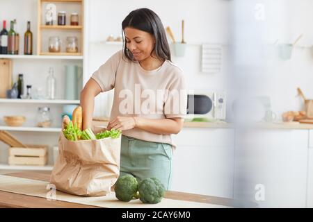 Portrait à la taille de l'élégante femme de course mixte déballant des sacs avec de la nourriture tout en se tenant dans la cuisine blanche intérieur et souriant, service de livraison de nourriture, espace de copie Banque D'Images