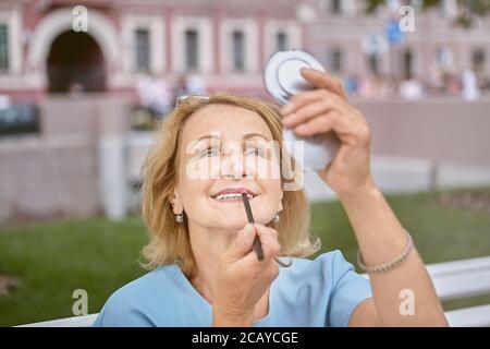 Femme blanche de race blanche âgée d'environ 60 ans, elle marche dans le centre-ville et se maquille avec un miroir de poche. Elle est active, gaie Banque D'Images