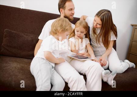 la jeune famille caucasienne était composée d'enfants, de petite fille, de fils et de parents assis sur un canapé portant des vêtements décontractés, en utilisant une tablette de gadget moderne Banque D'Images